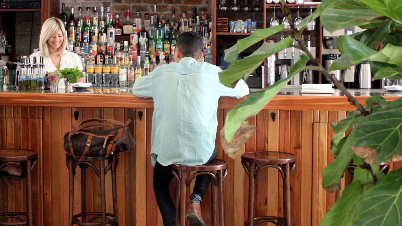 A man and woman at a wooden bar with bottles and stools.