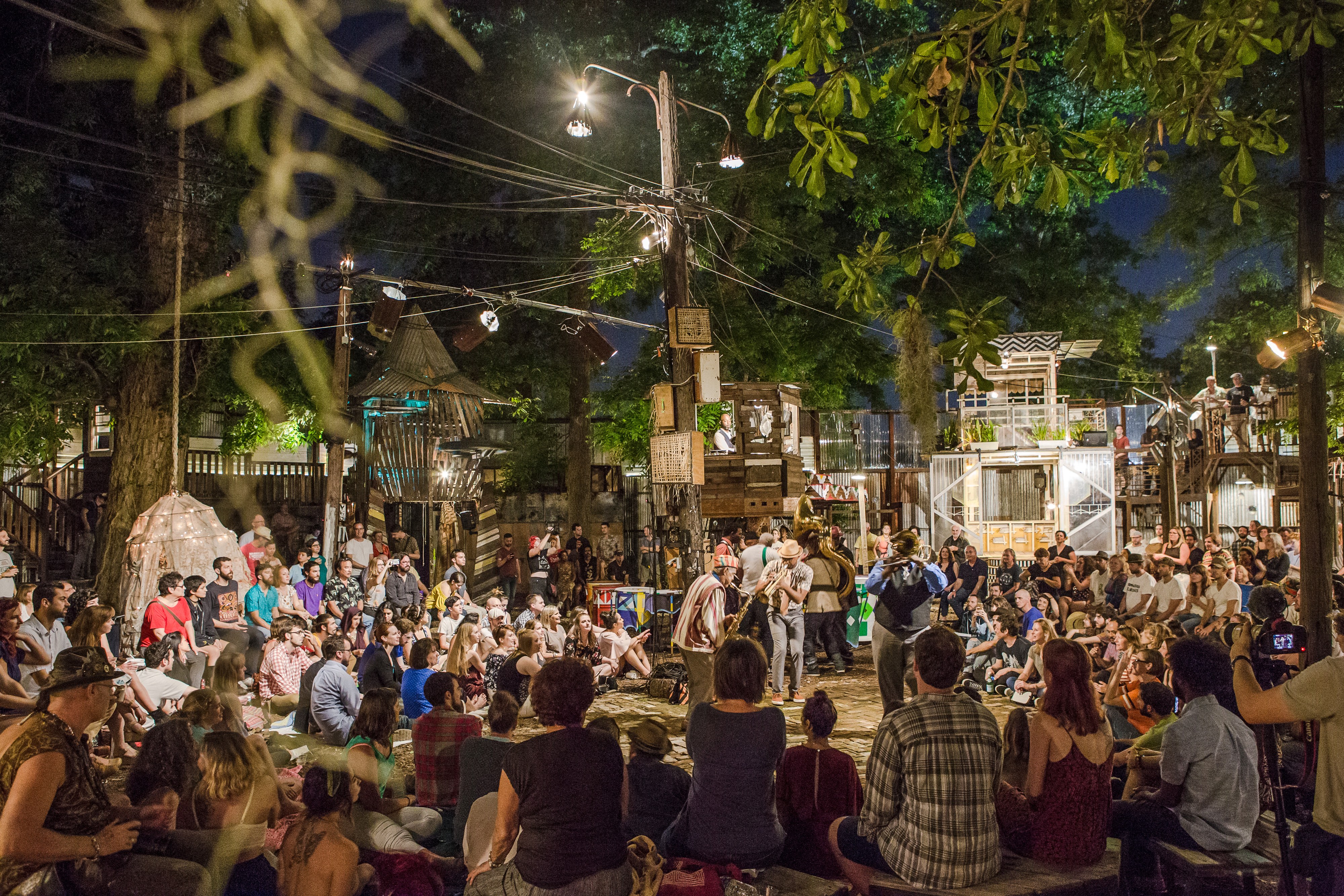A nighttime scene of a crowd gathered around a stage in an outdoor setting with trees and lights.